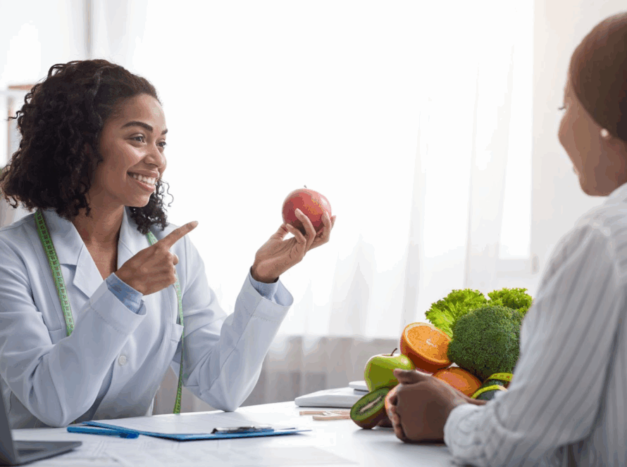A cheerful nutritionist gives counseling for nutrition, pointing to an apple while advising a patient on healthy eating.