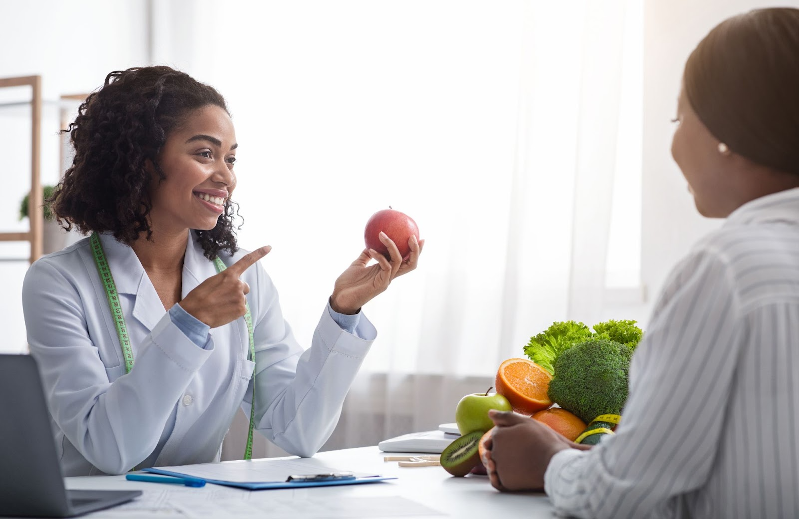 A cheerful nutritionist gives counseling for nutrition, pointing to an apple while advising a patient on healthy eating.
