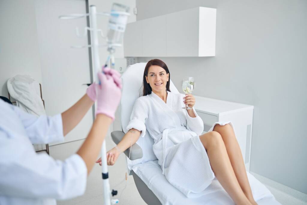 A woman holding a glass of water enjoys receiving intravenous therapy in a beauty clinic